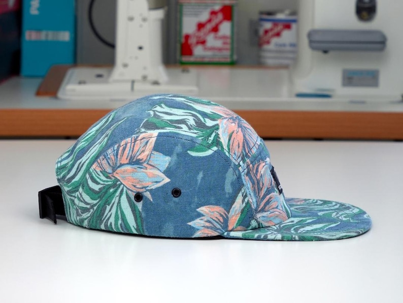 Pu&ograve; includere: Cappellino blu a cinque pannelli con visiera piatta, con un motivo floreale tropicale verde, rosa e bianco. Ha un cinturino regolabile nero e due occhielli neri. Un accessorio alla moda.