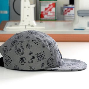 Pu&ograve; includere: Cappellino grigio a cinque pannelli con motivo paisley nero. Il cappellino ha una visiera piatta e un cinturino regolabile nero. Realizzato in tessuto morbido, &egrave; adatto per l'abbigliamento casual.