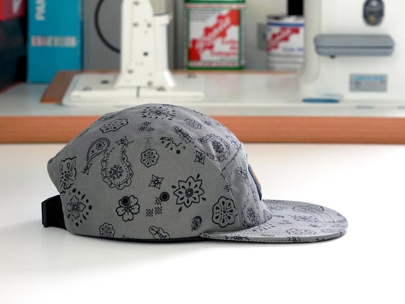 Pu&ograve; includere: Cappellino grigio a cinque pannelli con motivo paisley nero. Il cappellino ha una visiera piatta e un cinturino regolabile nero. Realizzato in tessuto morbido, &egrave; adatto per l'abbigliamento casual.