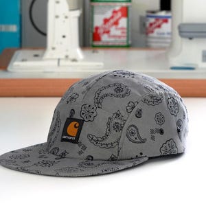 Pu&ograve; includere: Cappellino grigio a cinque pannelli con motivo paisley nero e visiera piatta. Il cappellino presenta il logo Carhartt sul davanti. Un accessorio elegante per un look casual.
