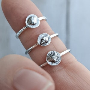 May include: Three silver rings featuring different seashell designs. The rings are stacked on a finger.