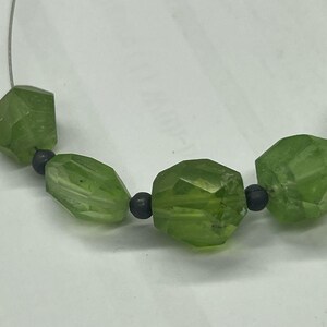 May include: A close-up of a necklace featuring faceted, light green beads. The beads are interspersed with small, dark gray beads. The necklace is strung on a thin, silver-colored wire, creating a simple and elegant design.