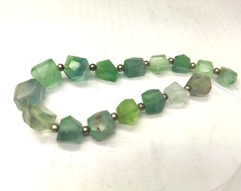 Op de afbeelding: Een ketting met onregelmatige, facetgeslepen kralen in groen- en blauwgroentinten. De kralen zijn geregen met kleine, zilverkleurige metalen kralen. De ketting wordt getoond tegen een witte achtergrond.