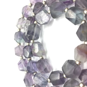 May include: A close-up of a necklace featuring faceted, multi-toned purple and gray fluorite beads. The beads are strung together with small silver-colored metal beads. The necklace is displayed against a white background.