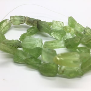 May include: A strand of light green peridot gemstone beads. The beads are irregularly shaped, with a rough, natural texture. The beads are strung on a thin, clear cord, and are suitable for jewelry making.
