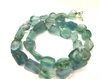 Op de afbeelding: Een ketting met onregelmatige, gepolijste fluorietstenen in blauwe en groene tinten. De stenen zijn geregen met een zilverkleurige sluiting. De ketting wordt gepresenteerd tegen een witte achtergrond, waardoor de natuurlijke kleuren en texturen van de edelstenen worden benadrukt.