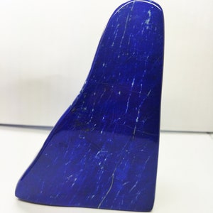 Puede incluir: Una escultura pulida de lapislázuli azul profundo con vetas blancas. La forma abstracta tiene una forma curva y asimétrica, con una superficie lisa y brillante. La escultura se presenta sobre un fondo blanco liso, resaltando su rico color y textura.