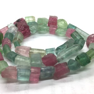 May include: A strand of raw tourmaline gemstones in various colors, including pink, green, and blue-green. The gemstones are irregularly shaped and appear to be uncut, with a natural, rough texture. The stones are strung together.