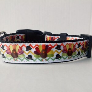 May include: White dog collar with a Thanksgiving turkey pattern. The collar has a black buckle and a silver D-ring.