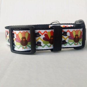 May include: A black dog collar with a white background and a repeating pattern of turkeys wearing pilgrim hats. The collar has a black buckle.