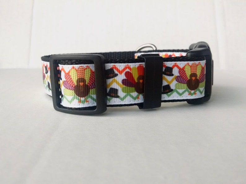 May include: A black dog collar with a white background and a repeating pattern of turkeys wearing pilgrim hats. The collar has a black buckle.