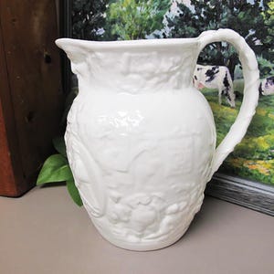 May include: White ceramic pitcher with a raised embossed design of a countryside scene. The pitcher has a handle that is shaped like a branch.