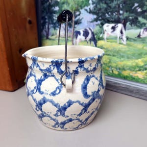 May include: A ceramic pitcher with a cream-colored base and a repeating blue sponge-painted pattern. The vessel features a small pouring spout, a rounded body, and a metal wire handle with a black cylindrical grip attached to two side loops.