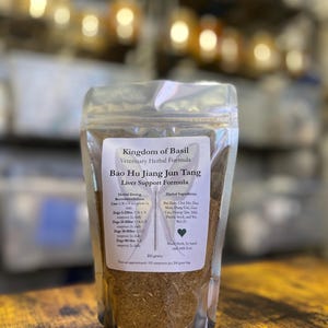 Puede incluir: Una bolsa resellable de polvo de hierbas marrón etiquetada como "Kingdom of Basil Veterinary Herbal Formula Bao Hu Jiang Jun Tang Liver Support Formula". La bolsa contiene 200 gramos de polvo e incluye una tabla de dosificación recomendada para gatos y perros.