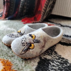 Puede incluir: Un par de zapatillas de fieltro gris con diseños de caras de búho. Los ojos del búho son amarillos y las zapatillas tienen una suela marrón. Las zapatillas están sobre una alfombra estampada.