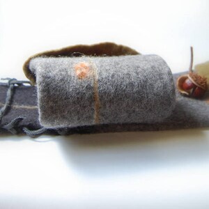 May include: A gray felt pouch with a brown felt flap and a brown felt acorn on top. The pouch is closed with a black leather strap with a decorative knot.