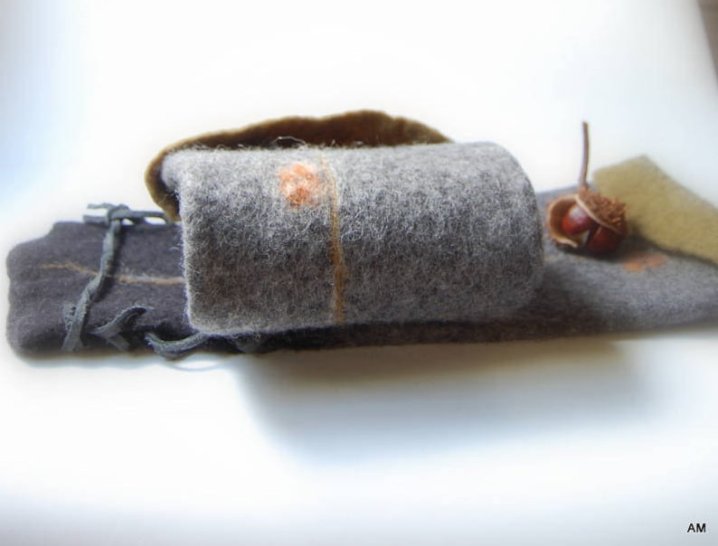 May include: A gray felt pouch with a brown felt flap and a brown felt acorn on top. The pouch is closed with a black leather strap with a decorative knot.