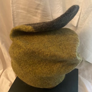 May include: A green felt hat with a gray felt brim. The hat is shaped like a rounded cone and has a unique, asymmetrical design.