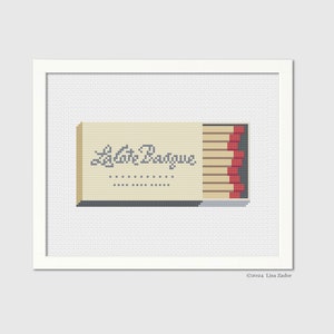 May include: Cross-stitch artwork of a matchbox with the text "La Cote Basque". The matchbox is beige with red-tipped matches visible. The artwork is framed in a white square frame.