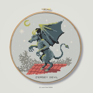 May include: Cross-stitch embroidery hoop featuring a gray, winged, and horned creature with a long tail, standing on a red brick rooftop. The creature is surrounded by a gray sky with a crescent moon and stars. The text "JERSEY DEVIL" is below the creature, and the text "(c) 2020 Lisa Zador" is below that.