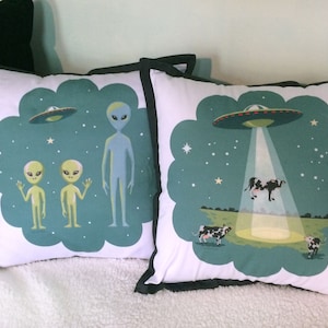 May include: Two throw pillows with a green background and white outlines. One pillow features two green aliens waving and a blue alien standing. The other pillow features a UFO with a beam of light shining down on a cow. The text on the pillow reads "Beam me up, Scotty!"