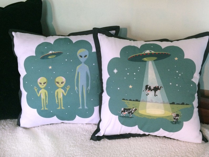 May include: Two throw pillows with a green background and white outlines. One pillow features two green aliens waving and a blue alien standing. The other pillow features a UFO with a beam of light shining down on a cow. The text on the pillow reads "Beam me up, Scotty!"
