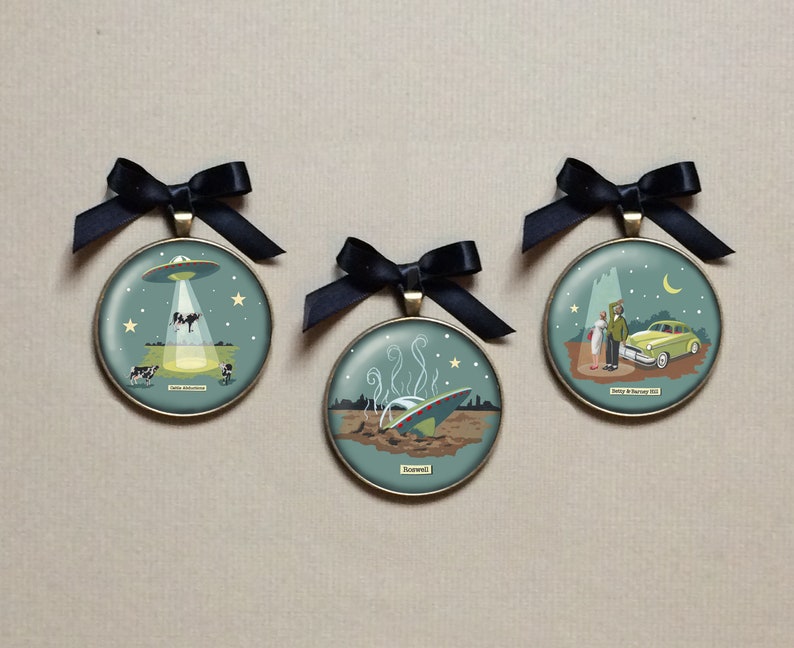 May include: Three round, gold-toned ornaments with black ribbon hangers. Each ornament features a different illustration of a UFO, with text that reads "Roswell", "Betty & Barney Hill", and "Cattle Mutilations".