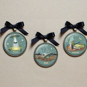 May include: Three round, gold-toned ornaments with black ribbon hangers. Each ornament features a different illustration of a UFO, with text that reads "Roswell", "Betty & Barney Hill", and "Cattle Mutilations".