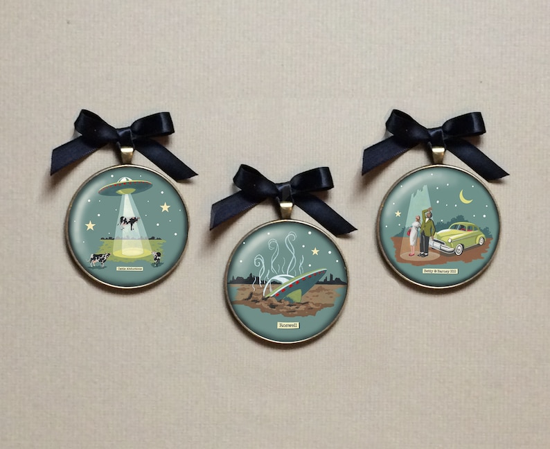 May include: Three round, gold-toned ornaments with black ribbon hangers. Each ornament features a different illustration of a UFO, with text that reads "Roswell", "Betty & Barney Hill", and "Cattle Mutilations".