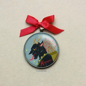 May include: A round, silver ornament with a blue background and a black and red illustration of Krampus, a mythical creature with horns, a long tongue, and a sack of twigs. The ornament has the text "Krampus" written in red cursive.