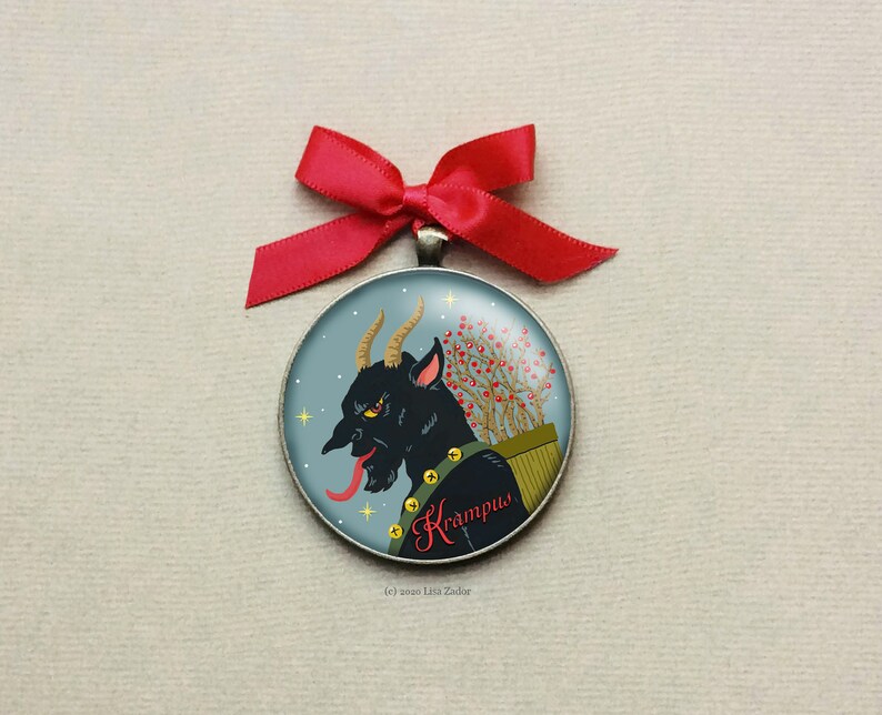 May include: A round, silver ornament with a blue background and a black and red illustration of Krampus, a mythical creature with horns, a long tongue, and a sack of twigs. The ornament has the text "Krampus" written in red cursive.