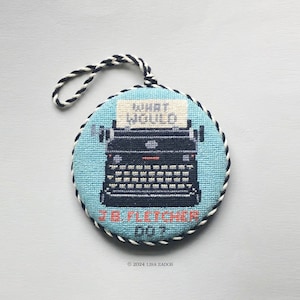 May include: A blue and white round needlepoint ornament with a black typewriter design. The typewriter has a white paper with the text "What Would J.B. Fletcher Do?"