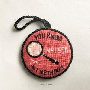 May include: A round, red needlepoint ornament with a black rope hanger. The ornament features a magnifying glass with the text "You Know Watson My Methods".