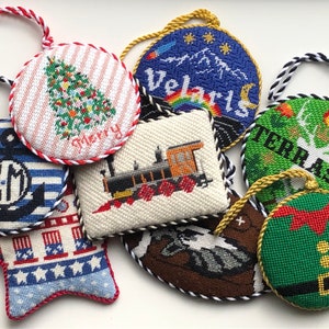 May include: Nine handmade needlepoint ornaments with various designs, including a Christmas tree, a train, a mountain scene, a deer, a gnome, and a nautical anchor. The ornaments are all round or star-shaped and have a red, white, and blue color scheme.