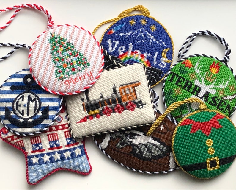 May include: Nine handmade needlepoint ornaments with various designs, including a Christmas tree, a train, a mountain scene, a deer, a gnome, and a nautical anchor. The ornaments are all round or star-shaped and have a red, white, and blue color scheme.