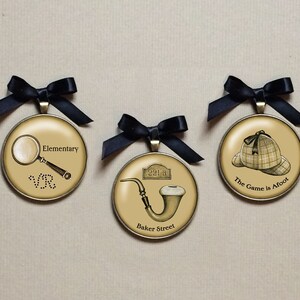 May include: Three round, gold-toned ornaments with black ribbon bows. The ornaments feature images related to Sherlock Holmes: a magnifying glass and the words "Elementary", a pipe and the words "Baker Street", and a deerstalker hat and the words "The Game is Afoot".