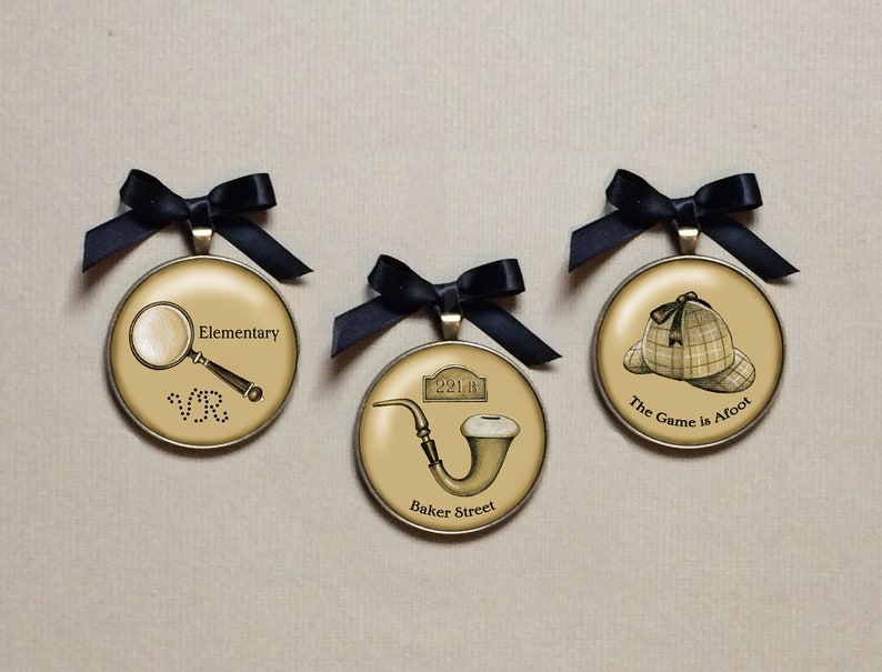 May include: Three round, gold-toned ornaments with black ribbon bows. The ornaments feature images related to Sherlock Holmes: a magnifying glass and the words "Elementary", a pipe and the words "Baker Street", and a deerstalker hat and the words "The Game is Afoot".