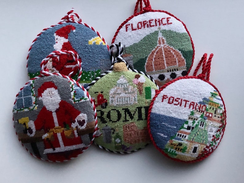 May include: Five round needlepoint ornaments with red and white trim. The ornaments feature scenes from Italy, including Santa Claus, the Colosseum, and the Duomo in Florence. The ornaments are labeled "ROMI", "FLORENCE", and "POSITANO".