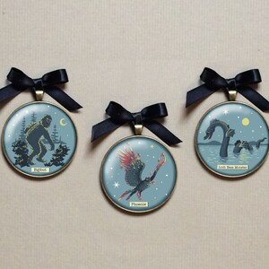 May include: Three round, gold-rimmed ornaments with blue backgrounds and black ribbon hangers. Each ornament features a different mythical creature: Bigfoot, the Loch Ness Monster, and the Phoenix. The ornaments are on a brown surface.