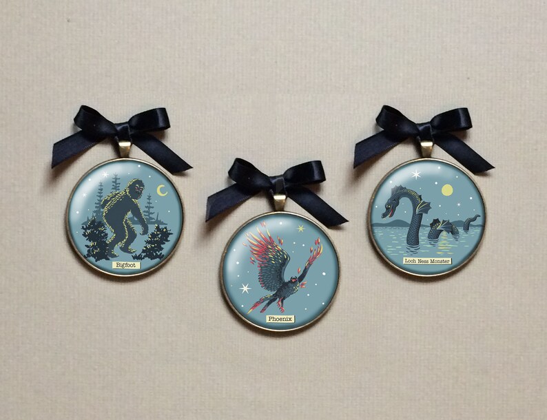 May include: Three round, gold-rimmed ornaments with blue backgrounds and black ribbon hangers. Each ornament features a different mythical creature: Bigfoot, the Loch Ness Monster, and the Phoenix. The ornaments are on a brown surface.