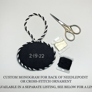 May include: Black and white oval needlepoint ornament with a white rope hanger and the date "2-19-22" embroidered on the front.  The ornament is accompanied by two spools of embroidery floss, one white and one black, and a pair of small gold-handled scissors.