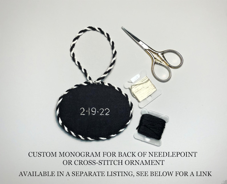 May include: Black and white oval needlepoint ornament with a white rope hanger and the date "2-19-22" embroidered on the front.  The ornament is accompanied by two spools of embroidery floss, one white and one black, and a pair of small gold-handled scissors.