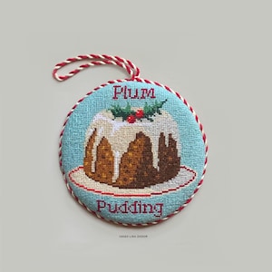 May include: A round, blue and white Christmas ornament with a red and white string for hanging. The ornament features a stitched image of a Christmas pudding with white icing and red berries on top. The text "Plum Pudding" is stitched in red on the ornament.