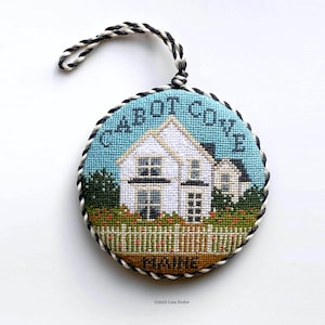 May include: A white house with a black roof and a white picket fence, surrounded by green bushes and red flowers. The house is on a blue background with the text "CABOT COVE MAINE" in black. The image is a round needlepoint ornament with a black and white braided cord.