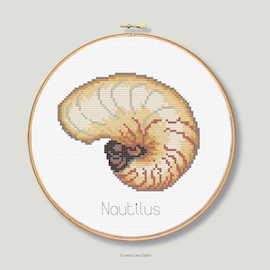 May include: A cross-stitch pattern of a nautilus shell in a circular embroidery hoop. The shell is a light beige color with a darker brown center. The text "Nautilus" is printed below the shell.