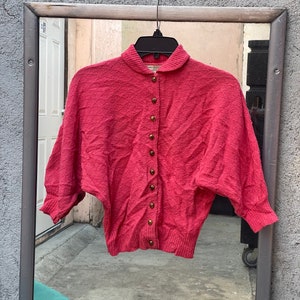 May include: A pink knitted cardigan with a button-up front and a Peter Pan collar. The cardigan has a textured knit and is cropped.