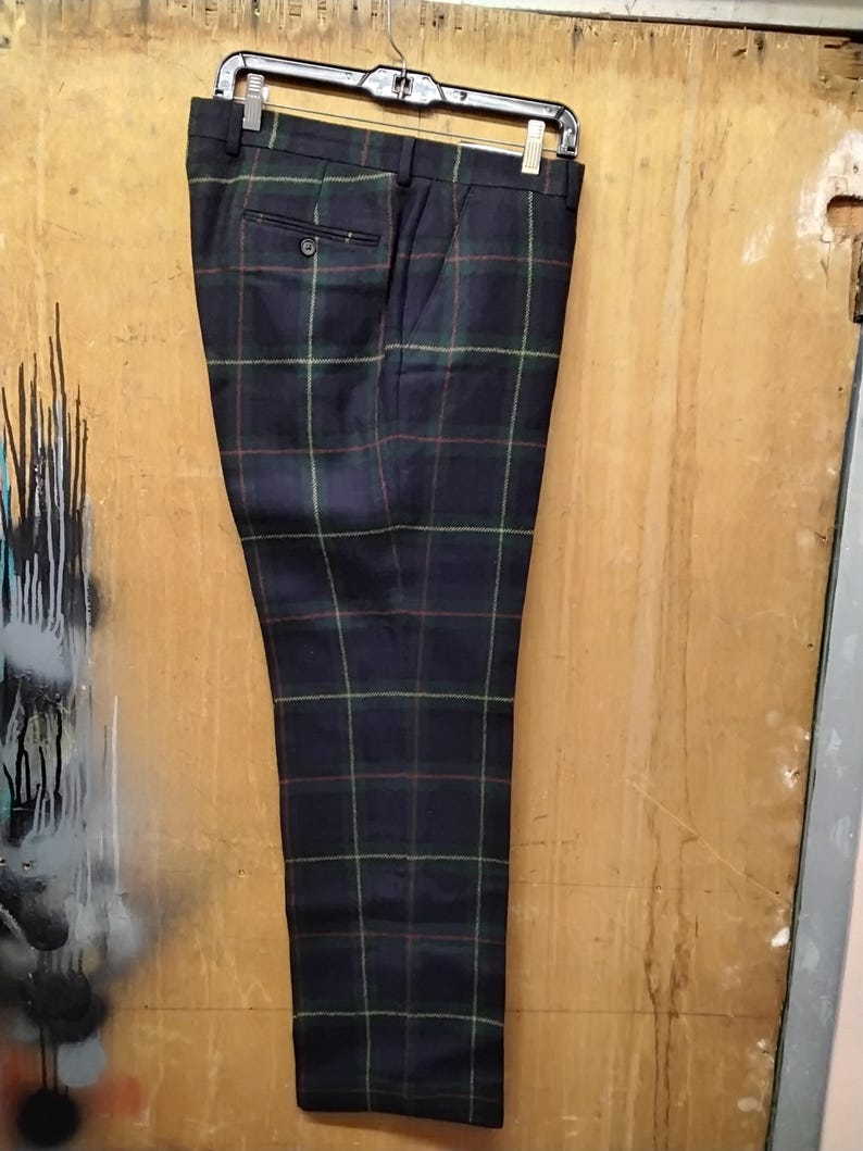 May include: Navy blue plaid trousers hanging on a black hanger. The trousers feature a classic plaid pattern in green, red, and yellow lines over a navy background. The pants have a buttoned back pocket and a button closure.