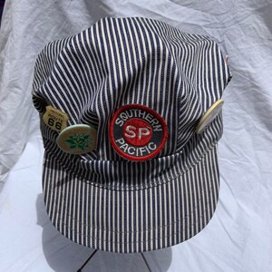 May include: A blue and white striped railway engineer's cap with a Southern Pacific logo patch and various pins, including a "Historic Route 66" pin.
