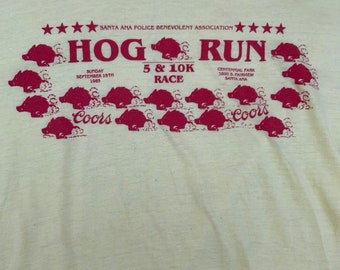 Könnte beinhalten: Ein Vintage-T-Shirt in Weiß mit einem roten Aufdruck von laufenden Schweinen. Der Text lautet "HOG RUN", "Sonntag, 15. September 1965", "5 & 10K RACE", "Centennial Park 3000 S. Fairview Santa Ana" und "Coors".