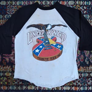 May include: White and black raglan-style t-shirt featuring a guitar design with the words "Lynyrd Skynyrd" and "Tribute Tour 1987". The guitar design incorporates red, white, and blue elements, with an eagle above the band's name.